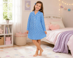 Cheerful young girl standing barefoot beside a pastel bed while wearing a blue gingham sleepshirt with long sleeves, collar neckline, and button-front styling. The gingham sleepshirt features a bright blue-and-white evenly spaced check pattern, lightweight breathable fabric, relaxed above-knee sleepshirt fit, cozy pajama comfort, and classic long-sleeve sleepwear styling, photographed with pastel bedroom décor, fairy lights, soft blankets, and bright natural indoor lighting