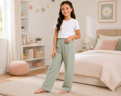 Young girl modeling sage green muslin pants with a white t-shirt in a softly decorated bedroom. The breathable muslin pants feature a comfortable loose fit, elastic waistband, and subtle textured fabric, photographed in a calm pastel interior with cozy bedding, decorative shelves, and natural daylight