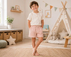 Happy young boy standing barefoot in a warm neutral-toned playroom while wearing blush pink muslin shorts with a cream henley t-shirt. The muslin shorts feature soft breathable double-layer muslin fabric, relaxed fit, elastic drawstring waistband, lightweight comfort, and casual lounge styling, photographed with teepee décor, woven textures, soft natural lighting, and cozy indoor surroundings