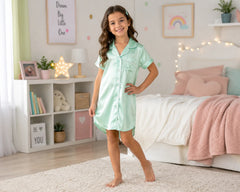 Smiling young girl standing in a softly decorated pastel bedroom while wearing a mint green satin sleepshirt with white piping, short sleeves, button-front pajama styling, chest pocket detail, and relaxed above-knee silhouette. The satin sleepshirt features glossy satin texture, lightweight silky fabric, elegant sleepwear comfort, premium smooth finish, and soft luxury pajama styling, photographed with pastel bedding, fairy lights, plush décor accents, and bright cozy bedroom atmosphere