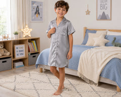 Happy young boy posing barefoot in a modern bedroom while wearing a silver grey satin sleepshirt with white piping, short sleeves, button-front pajama styling, chest pocket detail, and relaxed above-knee fit. The satin sleepshirt features shiny satin fabric, smooth silky finish, soft lightweight comfort, premium pajama-inspired tailoring, and elegant sleepwear styling, photographed with blue bedding, cozy rug texture, illuminated star décor, and stylish kids bedroom interior