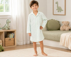 Happy young boy posing barefoot in a neutral-toned bedroom while wearing a soft sage striped seersucker sleepshirt with long sleeves, button-front design, classic pajama collar, chest pocket detail, and relaxed above-knee fit. The seersucker sleepshirt features lightweight textured fabric, breathable puckered construction, comfortable sleepwear styling, soft natural drape, and premium casual nightwear appearance, photographed with dinosaur-themed décor, plush toys, woven storage accents, and a bright family