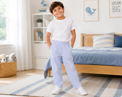Young boy standing in a nautical-themed kids bedroom wearing blue striped seersucker pants with a white t-shirt and white sneakers. The breathable seersucker pants feature vertical blue-and-white stripes, elastic waistband, and relaxed everyday comfort fit, styled in a bright bedroom with blue bedding, soft rugs, and minimal coastal-inspired décor