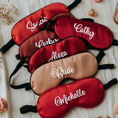 Set of personalized red and brown sleep masks with names embroidered on them, placed on a textured surface.