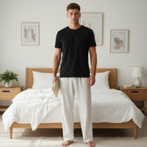 Man wearing a black t-shirt and white pants standing in a bedroom with a bed and nightstands.