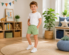Young boy smiling in a bright modern kids bedroom while wearing sage green cotton shorts paired with a plain white t-shirt and white sneakers. The soft cotton shorts feature a comfortable elastic waistband, breathable lightweight fabric, relaxed everyday fit, and casual summer styling, photographed with playful nursery décor, woven storage baskets, indoor plants, and warm natural sunlight