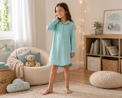 Happy young girl posing in a softly lit pastel bedroom while wearing a sky blue cotton sleepshirt with long sleeves and front button placket. The cotton sleepshirt features breathable cotton fabric, relaxed sleepshirt silhouette, lightweight soft texture, cozy loungewear comfort, and classic pajama-inspired styling, photographed with fairy lights, plush seating, soft pastel décor, and calming bedroom interiors