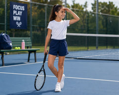 Active young girl standing on an outdoor tennis court wearing navy blue cotton sports shorts with a plain white t-shirt and white sneakers while holding a tennis racket. The sporty cotton shorts feature lightweight breathable fabric, elastic drawstring waistband, relaxed athletic fit, and comfortable movement-friendly styling, photographed in a sunny outdoor sports setting with tennis court fencing and motivational décor.