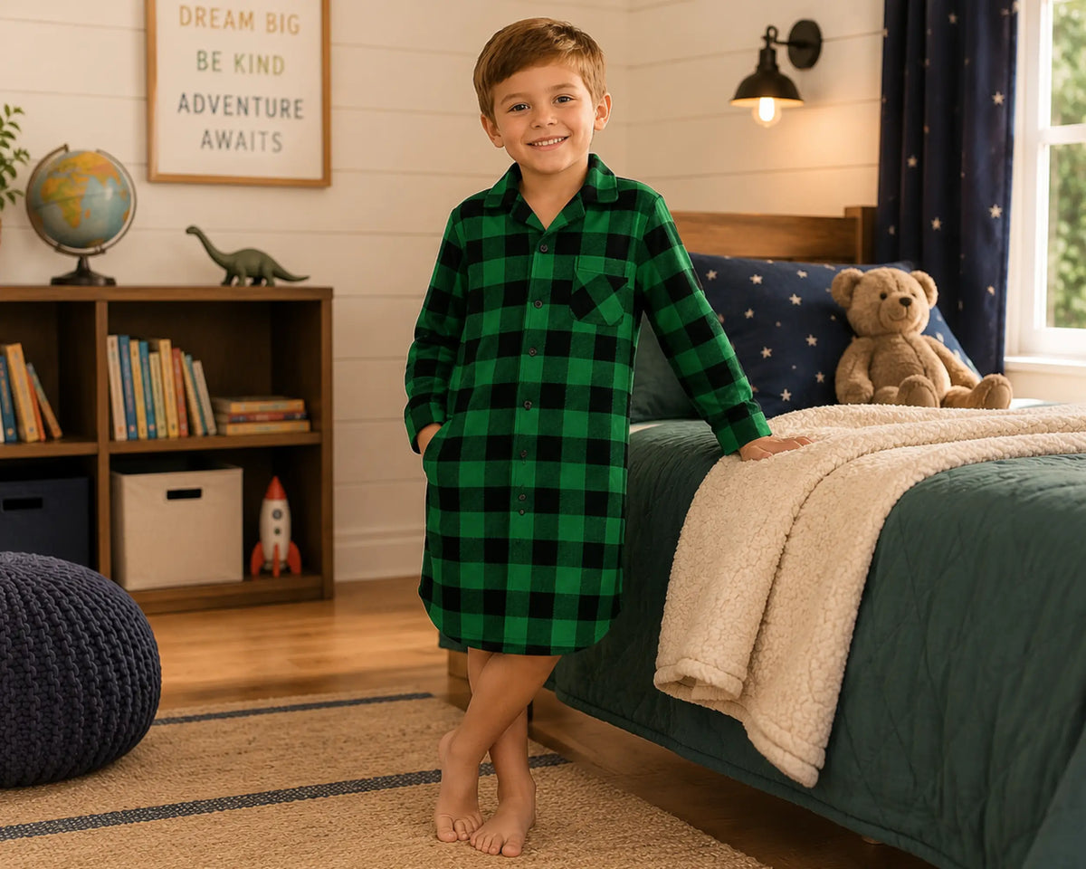 Happy young boy posing beside a cozy bed while wearing a green and black plaid flannel sleepshirt with long sleeves and button-front collar design. The flannel sleepshirt features a bold dark plaid pattern, soft brushed flannel fabric, relaxed oversized sleepshirt silhouette, warm cozy sleepwear comfort, and classic lumberjack-inspired styling, photographed with wooden bedroom décor, plush blankets, dinosaur accents, warm indoor lighting, and cozy nighttime aesthetics