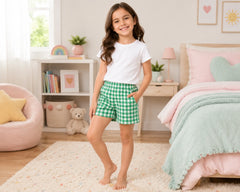 Smiling young girl posing in a pastel-themed kids bedroom while wearing green and white gingham shorts paired with a plain white t-shirt. The gingham shorts feature an evenly spaced check pattern, elastic waistband, lightweight cotton fabric, relaxed casual fit, and soft breathable comfort, photographed with cozy bedroom décor, pastel cushions, soft rugs, and bright natural daylight