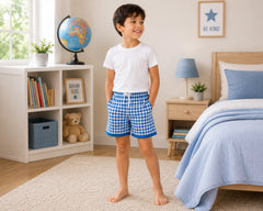 Happy young girl standing barefoot in a modern kids bedroom while wearing red and white gingham shorts with a plain white t-shirt. The gingham shorts feature a classic evenly aligned check pattern, lightweight breathable fabric, elastic waistband, comfortable relaxed fit, and playful everyday styling, photographed with warm indoor lighting, soft pastel décor, storage shelves, and cozy bedroom furniture