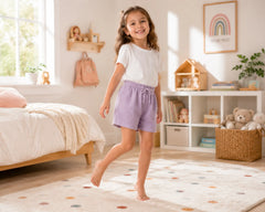 Smiling young girl walking barefoot in a soft pastel kids bedroom while wearing lavender muslin shorts paired with a plain white t-shirt. The muslin shorts feature lightweight breathable muslin fabric, soft textured finish, elastic drawstring waistband, relaxed casual fit, and airy everyday comfort, photographed with cozy pastel décor, natural sunlight, plush rugs, and modern bedroom styling