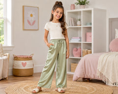Smiling young girl posing in a pastel bedroom wearing sage green satin pants with a cream short-sleeve top. The elegant satin pants feature a loose straight-leg fit, smooth shiny texture, adjustable drawstring waistband, and comfortable everyday styling, photographed in a soft pink-toned kids room with decorative shelves, woven rug, and cozy décor