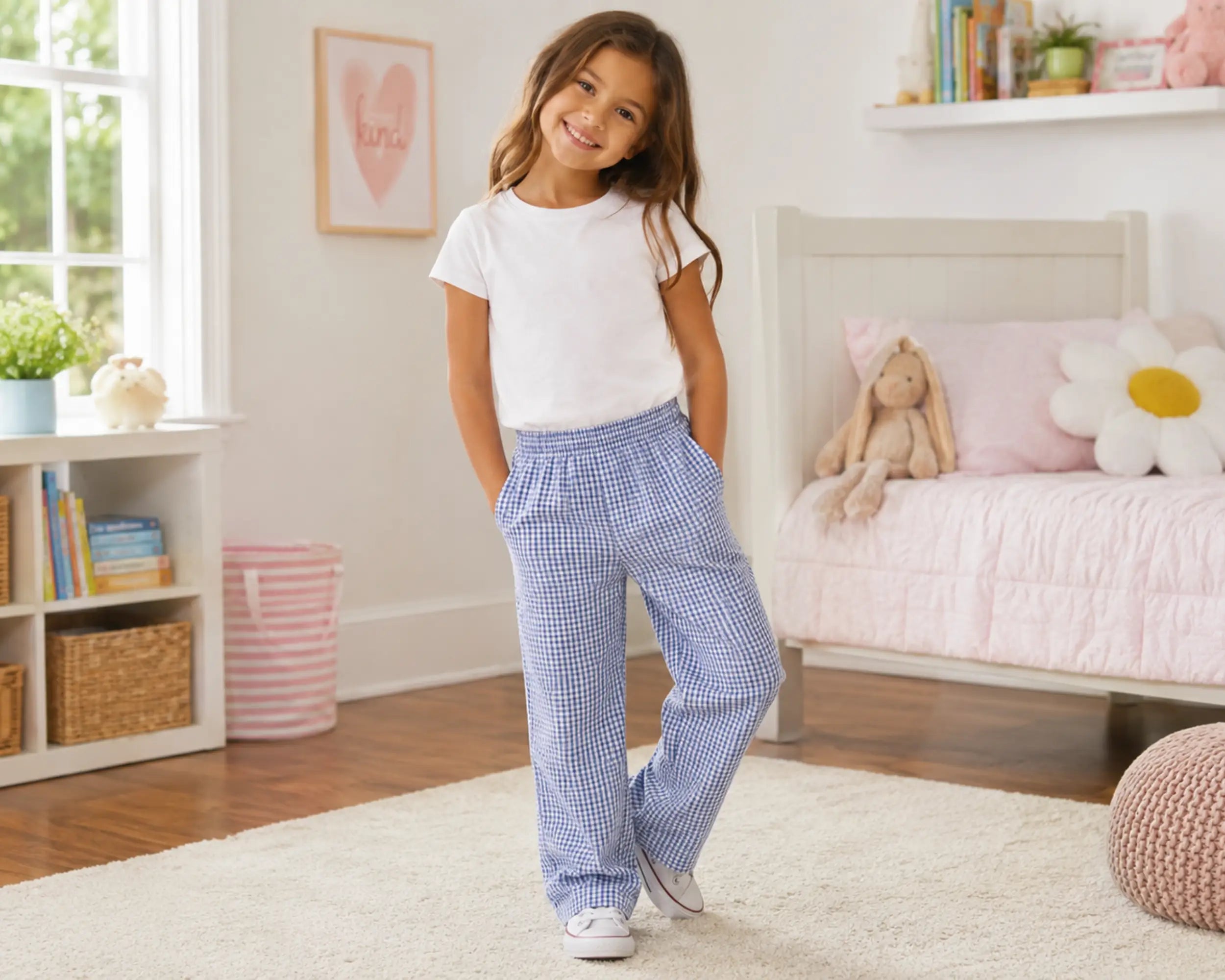 Young girl posing in a cheerful pastel bedroom while wearing blue gingham seersucker pants paired with a white t-shirt. The relaxed-fit pants feature a breathable lightweight texture, elastic waistband, and classic blue-and-white gingham check pattern, photographed in a soft kids bedroom with pink bedding, plush décor, and natural daylight