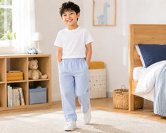 Smiling young boy walking barefoot in a bright modern bedroom wearing light blue seersucker pants with a plain white t-shirt. The lightweight striped seersucker pants feature a soft elastic waistband, breathable textured fabric, and relaxed straight-leg fit, styled in a cozy kids room with wooden furniture, soft rugs, and playful décor
