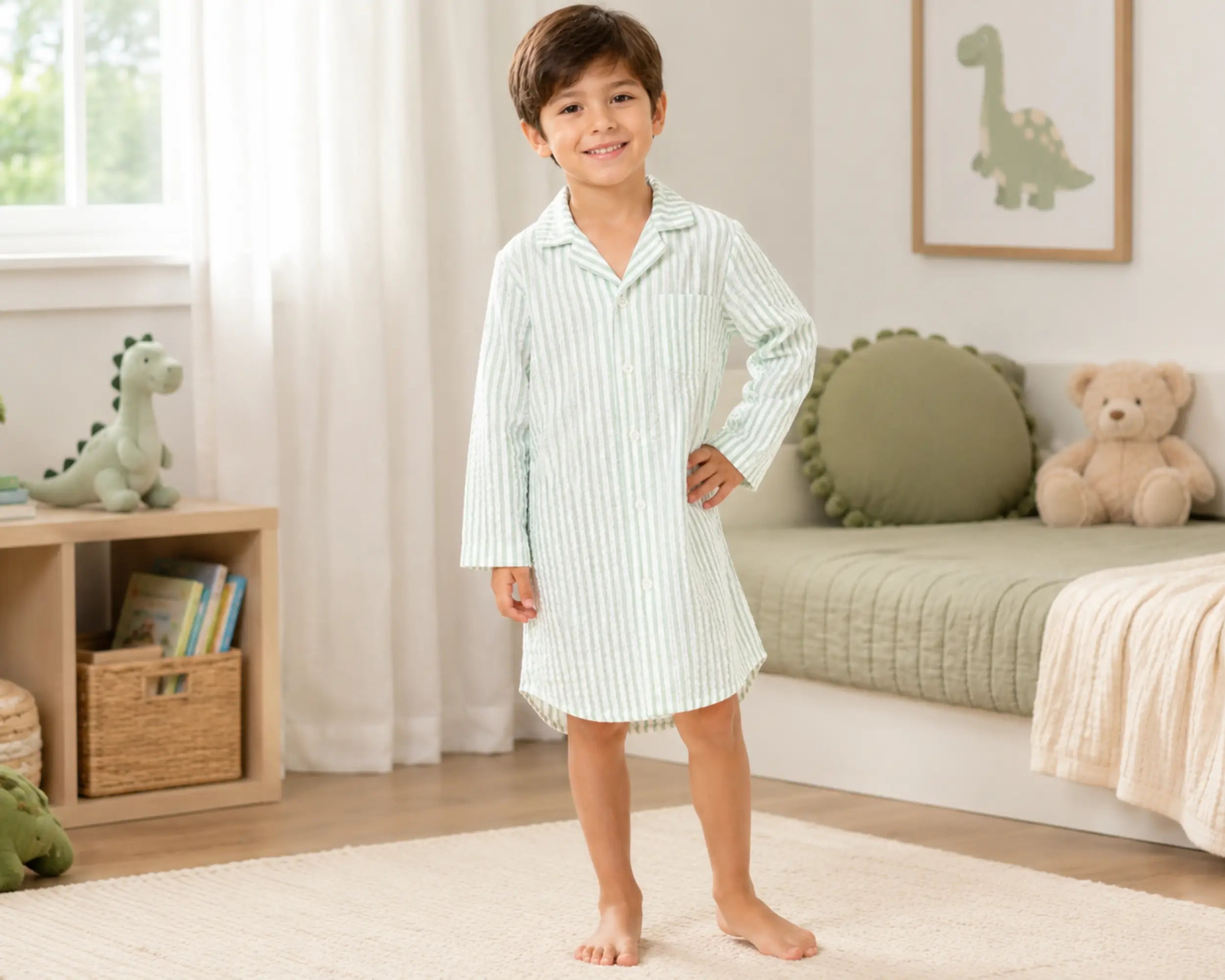 Happy young boy posing barefoot in a neutral-toned bedroom while wearing a soft sage striped seersucker sleepshirt with long sleeves, button-front design, classic pajama collar, chest pocket detail, and relaxed above-knee fit. The seersucker sleepshirt features lightweight textured fabric, breathable puckered construction, comfortable sleepwear styling, soft natural drape, and premium casual nightwear appearance, photographed with dinosaur-themed décor, plush toys, woven storage accents, and a bright family