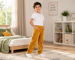 Smiling young boy posing confidently in a modern kids bedroom wearing mustard gold velvet pants with a plain white t-shirt and white sneakers. The premium velvet pants feature a rich velvety finish, comfortable elastic waistband, soft relaxed fit, and stylish everyday loungewear look, styled with wooden furniture, playful décor, and warm natural lighting