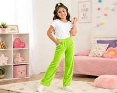 Cheerful young girl posing in a colorful kids bedroom while wearing bright neon green velvet pants paired with a white t-shirt and white sneakers. The vibrant velvet pants feature a smooth plush texture, elastic waistband, relaxed wide-leg fit, and playful comfortable styling, photographed in a bright pastel bedroom with colorful cushions, decorative lights, and fun kids décor