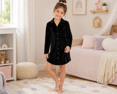 Smiling young girl standing barefoot in a cozy pastel bedroom while wearing a black velvet sleepshirt with long sleeves, button-front closure, classic collar, and relaxed above-knee silhouette. The velvet sleepshirt features soft plush velvet fabric, rich velvety texture, luxurious comfort, elegant sleepwear styling, and a smooth premium finish, photographed with blush bedding, decorative cushions, nursery décor, and a bright modern kids bedroom atmosphere