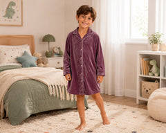 Happy young boy posing barefoot in a softly lit bedroom while wearing a dusty plum velvet sleepshirt with long sleeves, button-front design, classic pajama collar, and relaxed above-knee fit. The velvet sleepshirt features plush velvet fabric, smooth luxurious texture, comfortable warmth, premium sleepwear construction, and an elegant loungewear appearance, photographed with muted green bedding, teddy bear décor, soft natural lighting, and a cozy family-friendly bedroom setting
