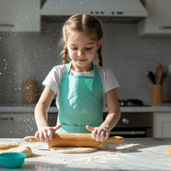 Child wearing a mint green apron using a wooden rolling pin to roll out dough in a kitchen