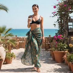 Woman in a black bikini top and leopard print sarong standing by a pool with ocean view.