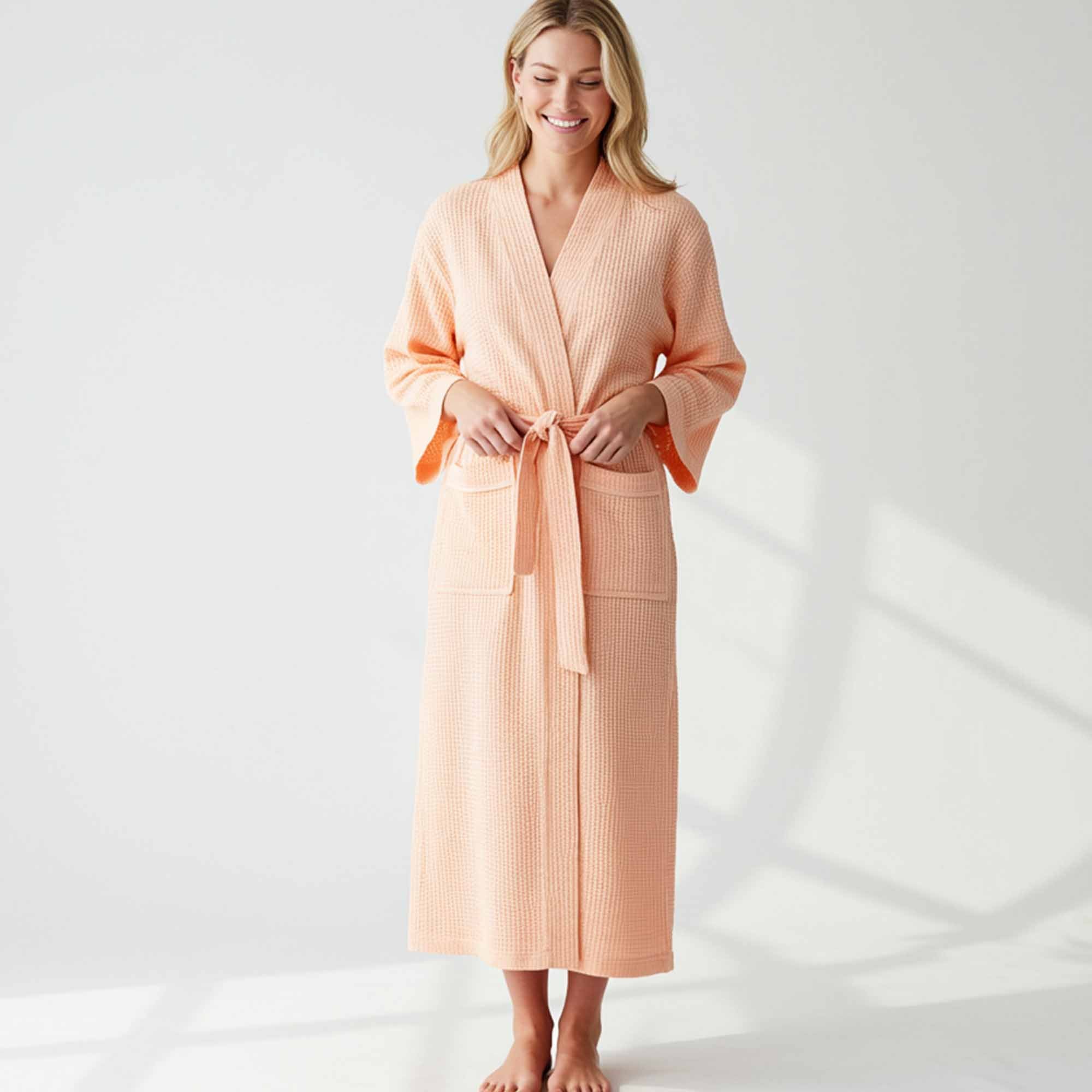wearing a long peach waffle-knit bathrobe with front pockets and a belt, standing in a bright minimal room.
