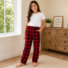 Young girl wearing red plaid pajama pants and a white t-shirt in a room with a wooden dresser and window.