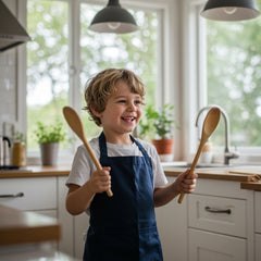 smiling toddler in cotton apron holding wooden spoons